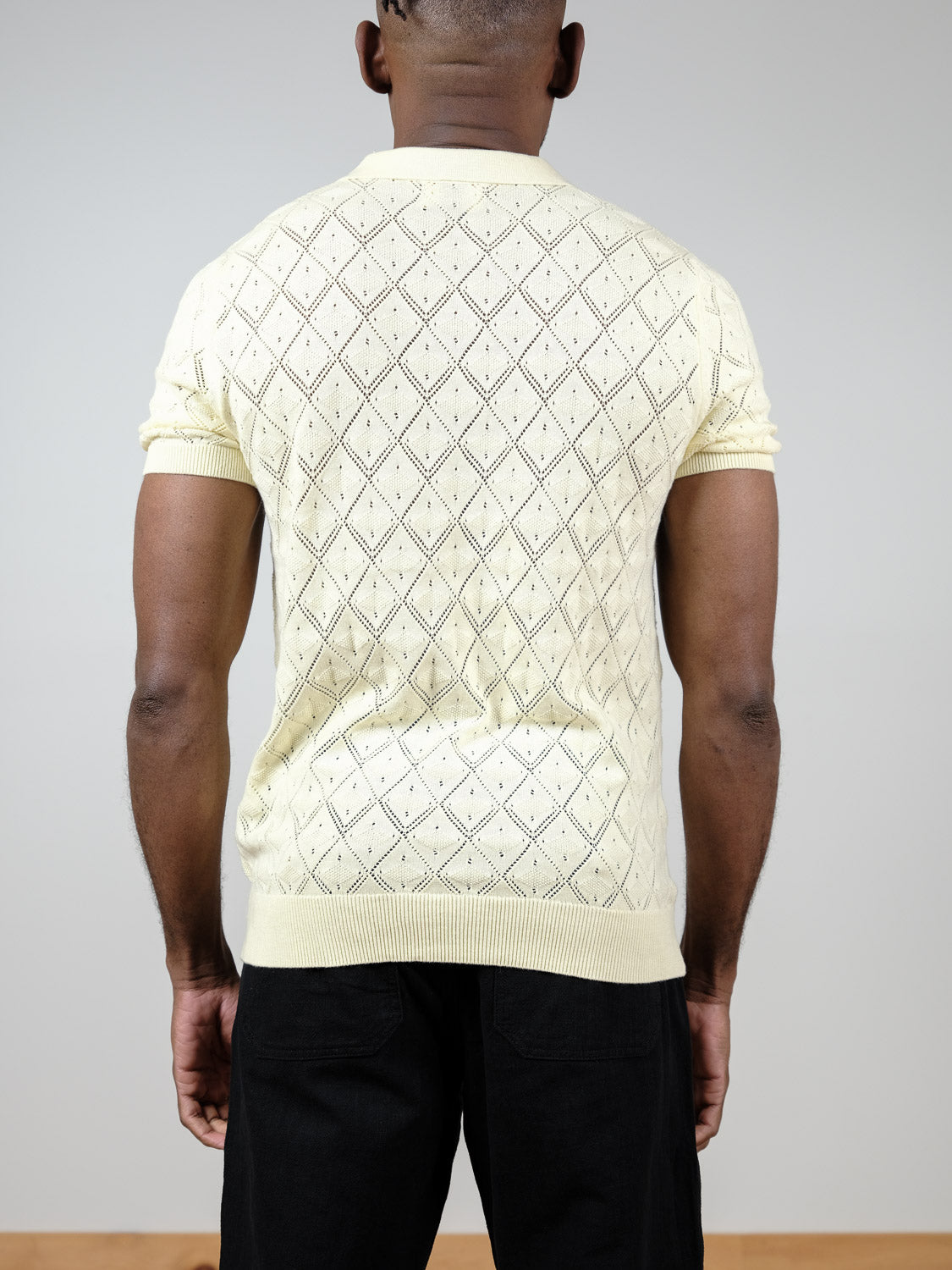 A person wearing the Agra Knitted Cotton Polo by ID Menswear, a short-sleeved light yellow sweater with a diamond pattern, paired with black pants, is shown from the back against a plain light gray background.