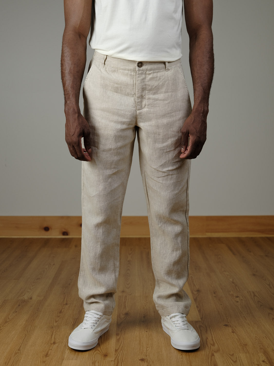 A person is indoors on a wooden floor, wearing a white t-shirt, white sneakers, and Casual Friday's Pandrup Linen Pants—beige summer pants with an elastic waistband. The photo shows the body from shoulders to feet.