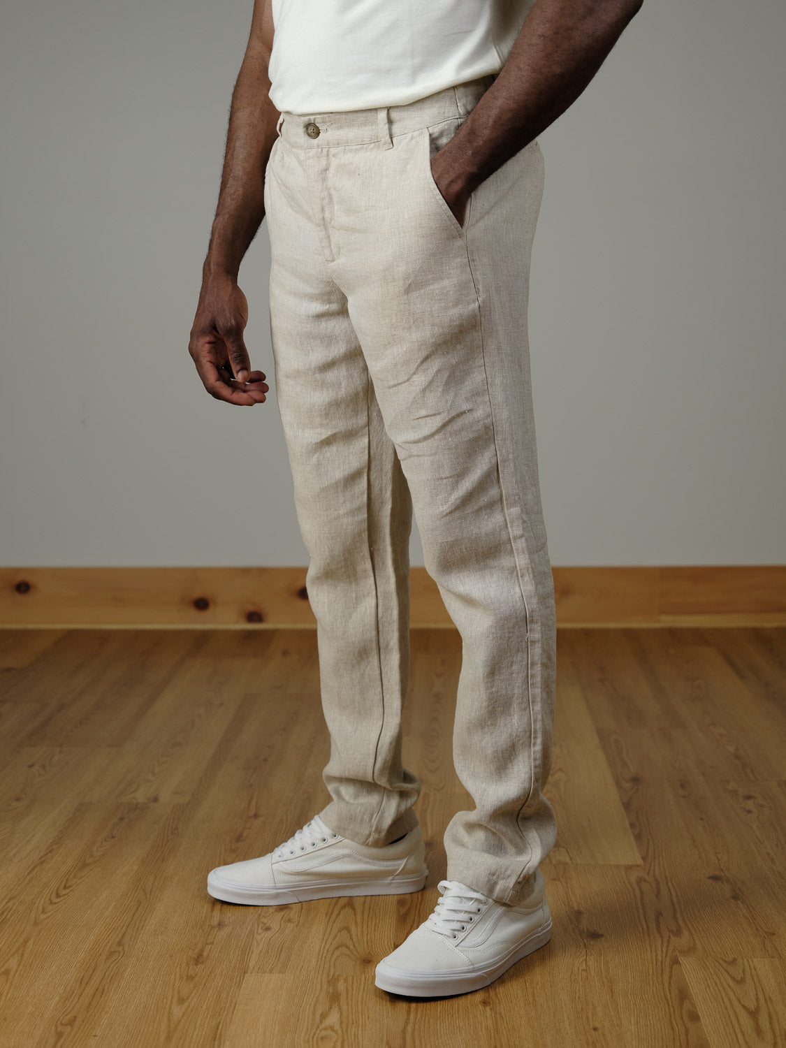 A person wearing Pandrup Linen Pants by Casual Friday, featuring an elastic waistband, stands on a wooden floor with hands in pockets. The pants are beige and styled with white sneakers; the upper body is mostly out of frame.