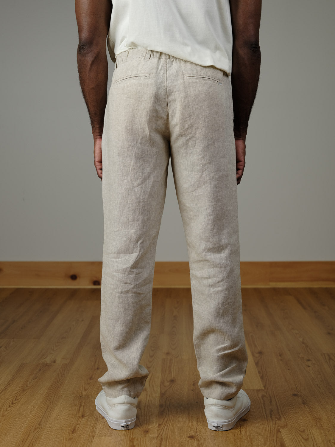 A person stands indoors on a wooden floor, facing away from the camera, wearing the Casual Friday Pandrup Linen Pants in light beige with an elastic waistband, paired with a white t-shirt and white sneakers.