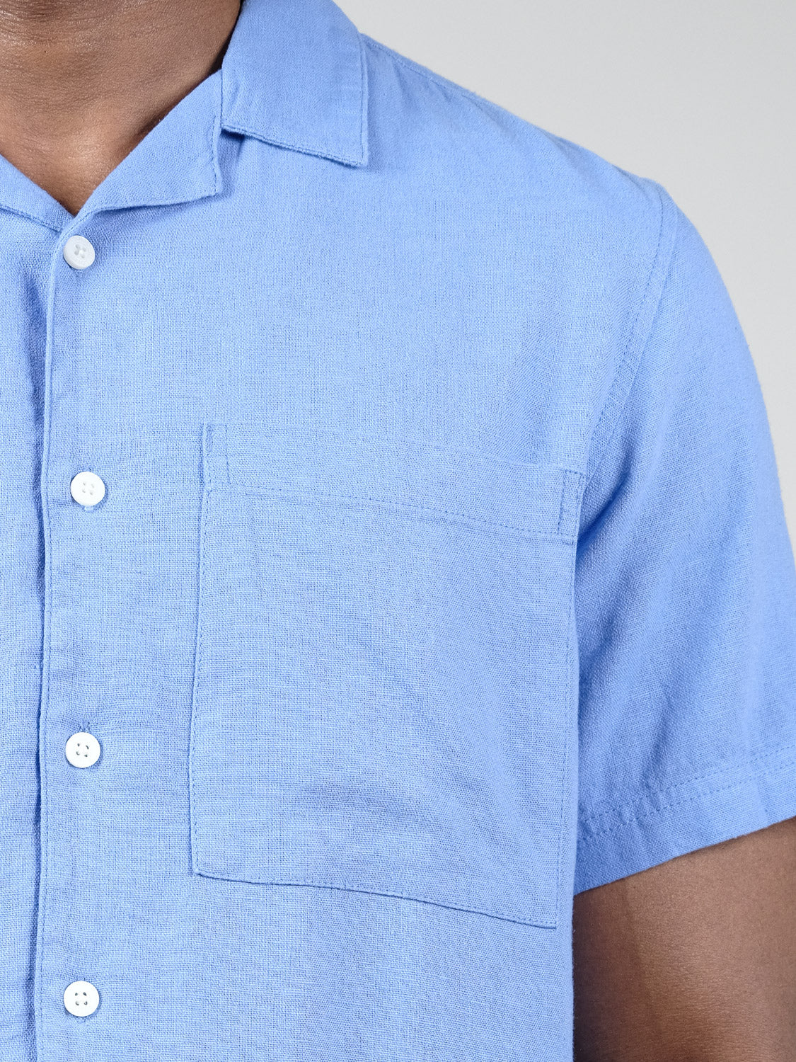 A close-up of a person wearing the Allan Cuba Woven Short Sleeve Shirt by !Solid, featuring light blue linen fabric, a relaxed collar, white buttons, and front chest pocket. Only the upper torso is visible.
