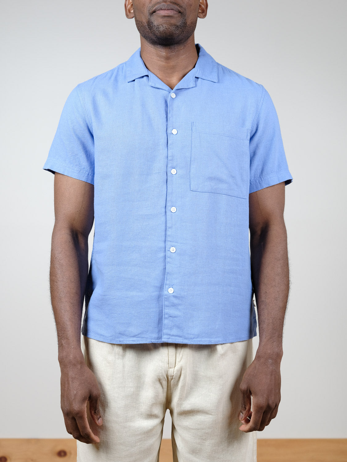 A man wears the !Solid Allan Cuba Woven Short Sleeve Shirt—a light blue linen button-up with a chest pocket—paired with light pants. He stands against a plain background, his face partially out of frame.