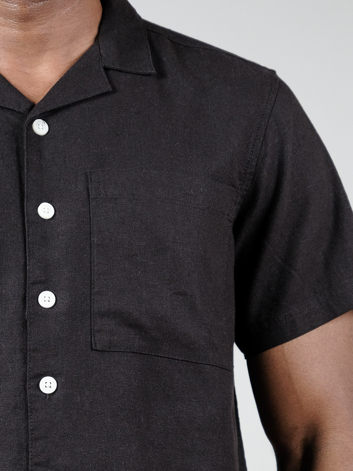 Close-up of a person wearing the Allan Cuba Woven Short Sleeve Shirt by !Solid—a black, short-sleeved button-up with white buttons and a chest pocket—showing only the upper torso against a plain background.