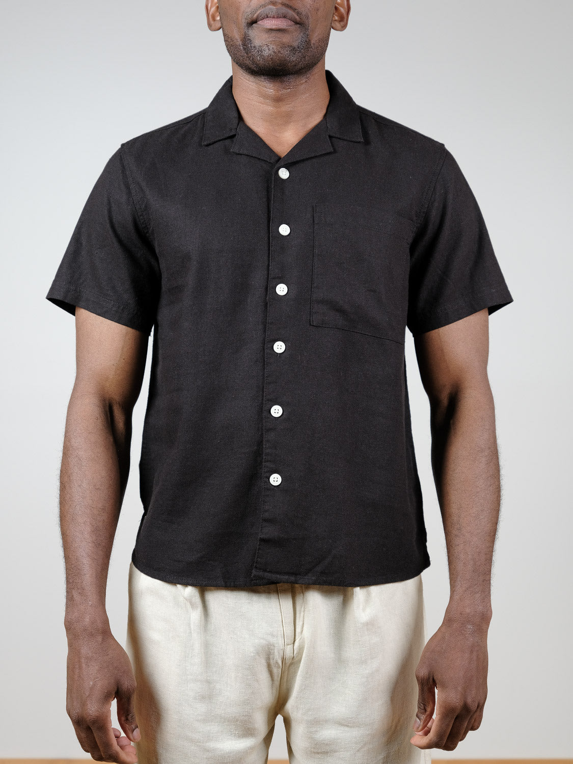 A man wears the !Solid Allan Cuba Woven Short Sleeve Shirt, a black button-up with a chest pocket, paired with cream pants. He stands against a plain light background, his face partially out of frame.