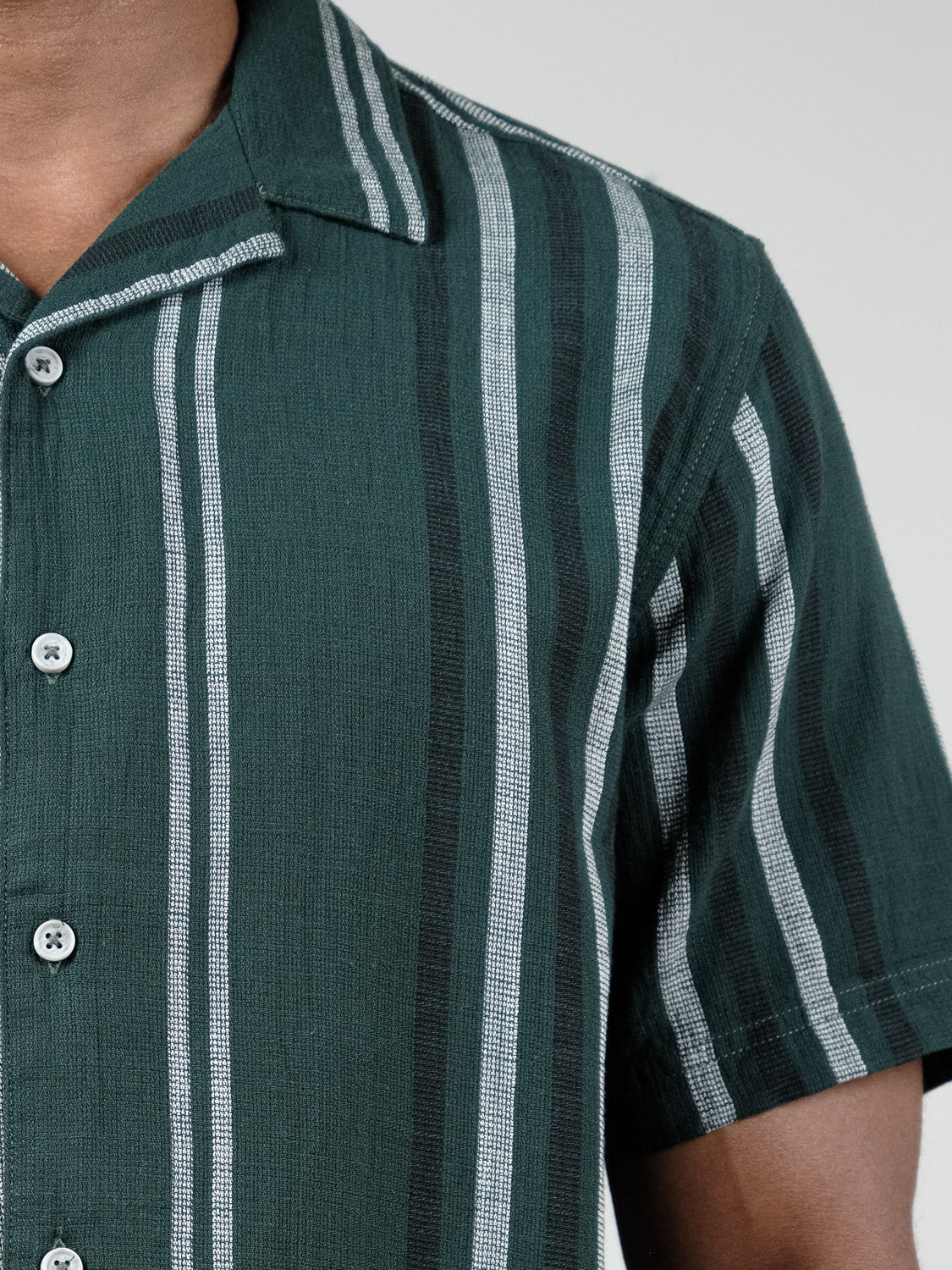 A close-up shows a person wearing the Isai Short Sleeve Shirt by !Solid, featuring green cotton fabric with vertical white and black stripes, highlighting the camp collar, upper chest, shoulder, and part of the arm.