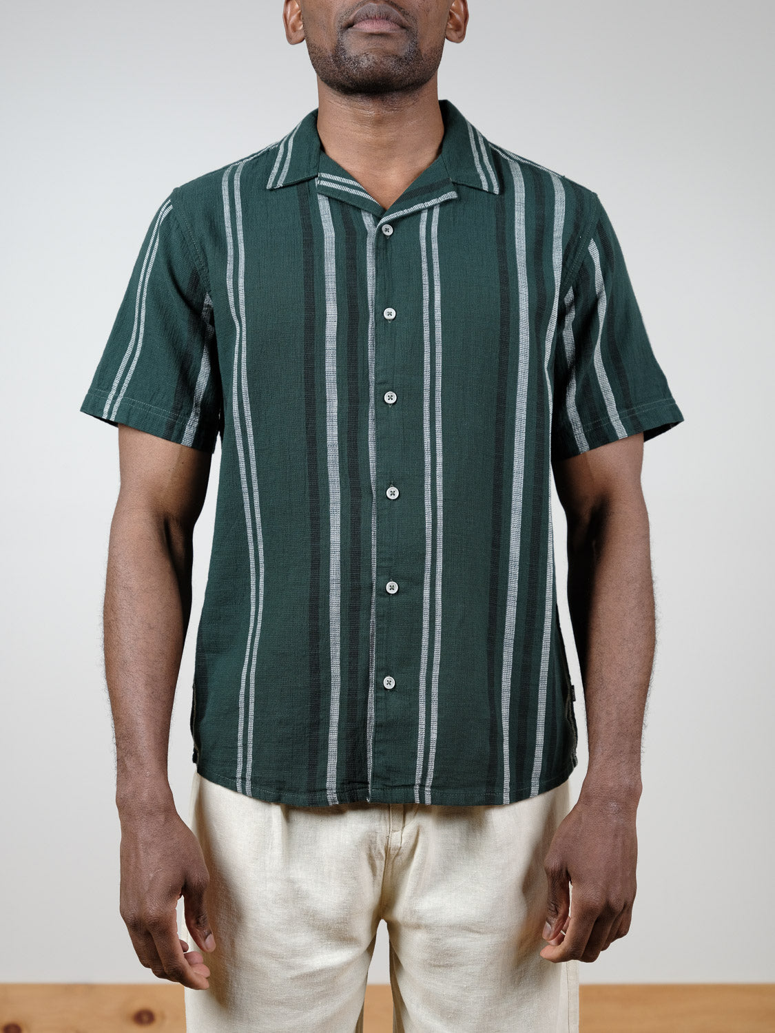 A man wears the !Solid Isai Short Sleeve Shirt—dark green with vertical white and gray stripes—paired with light-colored pants, standing against a plain light background.