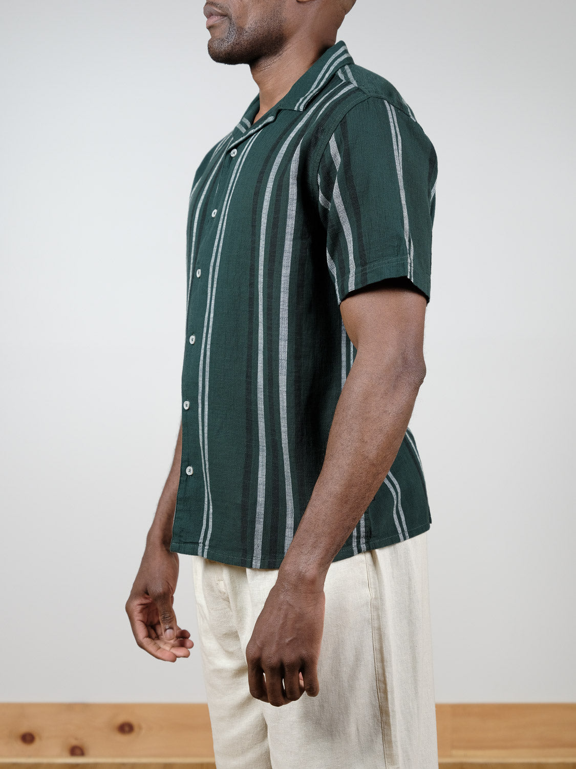 A man stands sideways wearing the Isai Short Sleeve Shirt by !Solid, a dark green camp collar shirt with white and gray vertical stripes, paired with light beige pants. The background is plain and uncluttered.