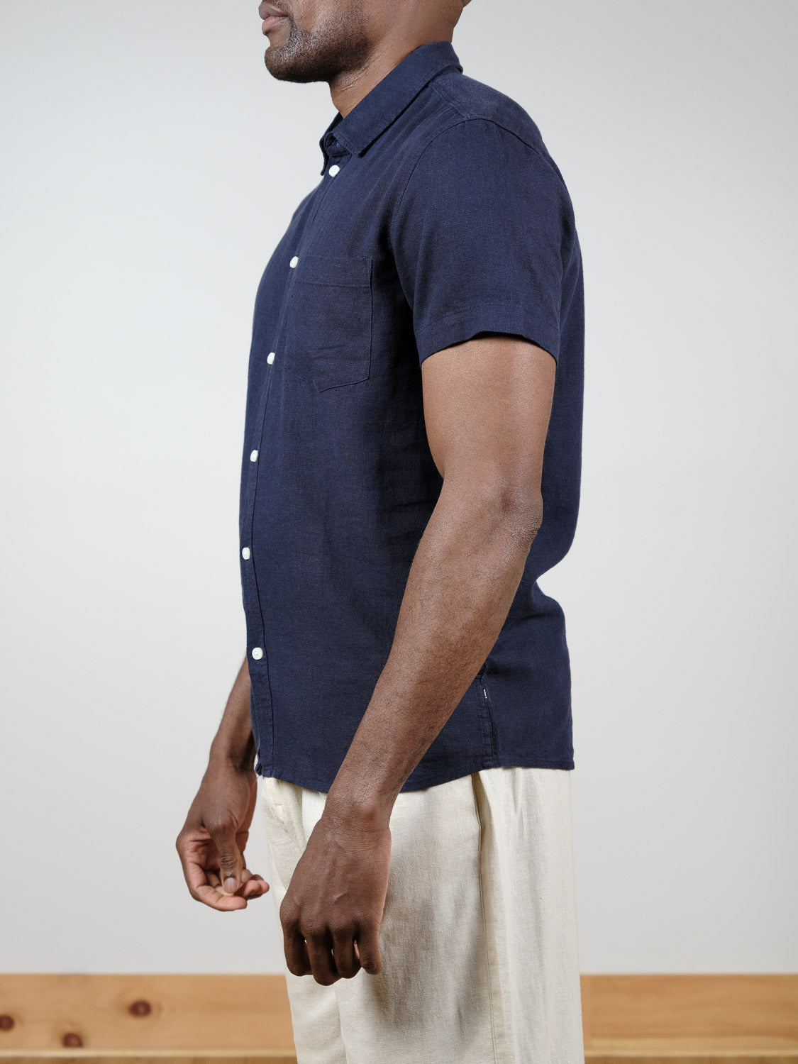 A person stands sideways against a plain light background with a wooden ledge, wearing the !Solid Allan Short Sleeve Shirt in navy blue paired with light beige pants.