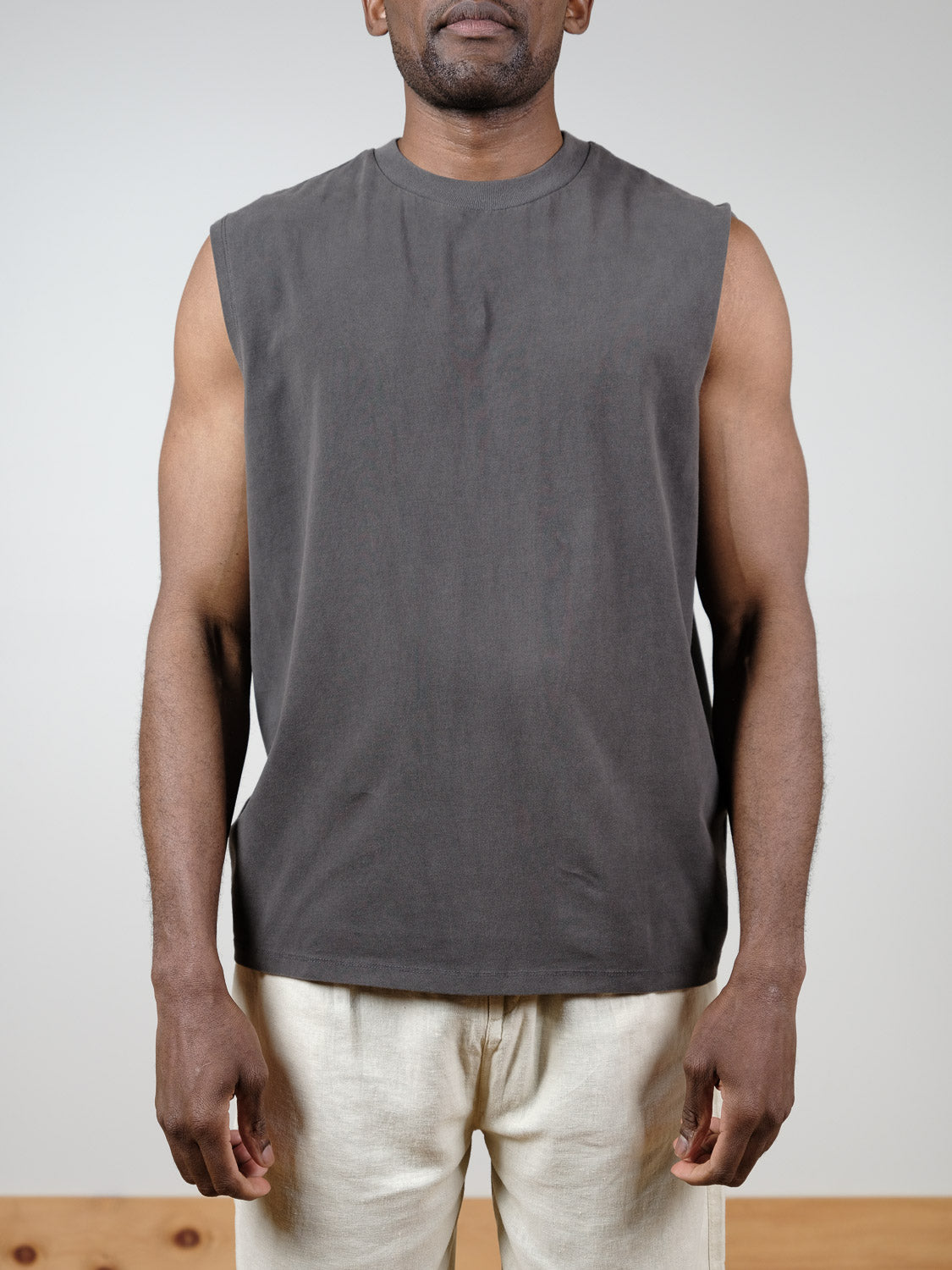 A man stands facing forward against a plain light background, wearing Industrie’s The Kosta Sleeveless Tee in dark gray and light beige pants, with his arms relaxed at his sides.