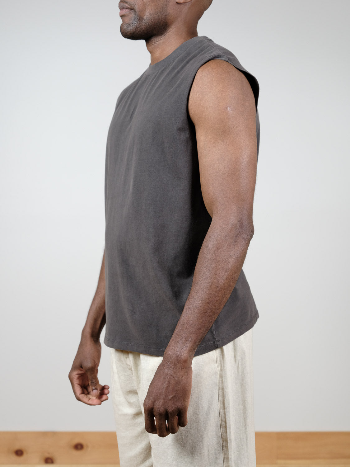A man stands in profile against a plain background, wearing Industrie’s The Kosta Sleeveless Tee in dark gray and light beige pants. His arms are relaxed by his sides, and his face is mostly out of frame.