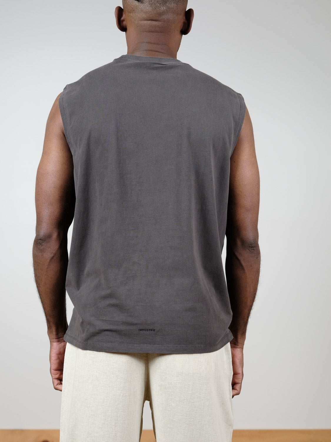 Someone stands facing away from the camera against a plain light background, wearing Industrie’s The Kosta Sleeveless Tee in dark gray and light beige pants.