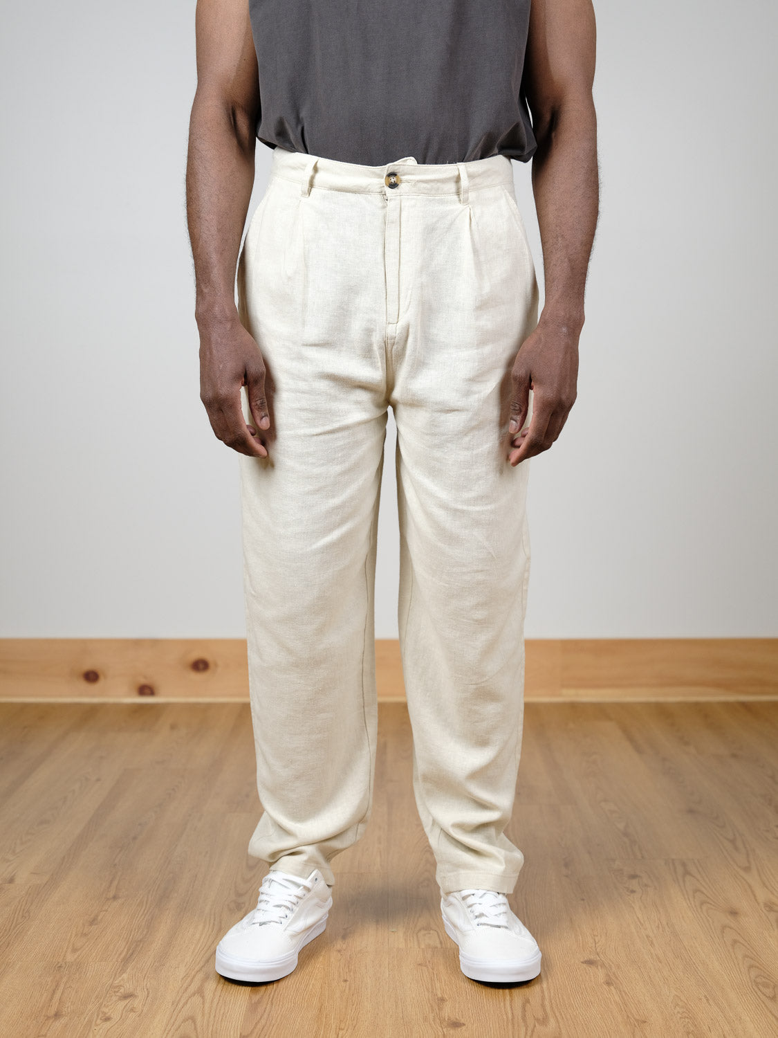 A person stands on a wooden floor in the !Solid Ethan Tapered Pleat Pant, a high-waisted light beige linen viscose blend with a tapered pleated design, paired with a dark gray shirt and white sneakers against a minimal plain background.