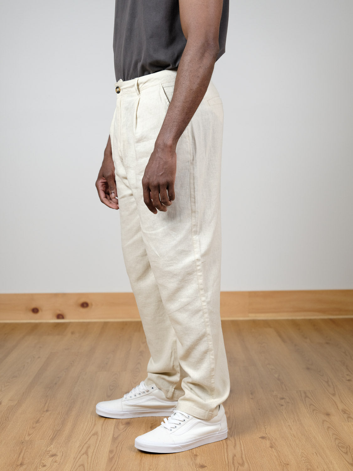 A person stands indoors on a wooden floor, wearing !Solid's Ethan Tapered Pleat Pant in light beige linen-viscose, paired with a dark shirt and white sneakers. The side-view photo captures them from shoulders to feet, arms relaxed at their sides.