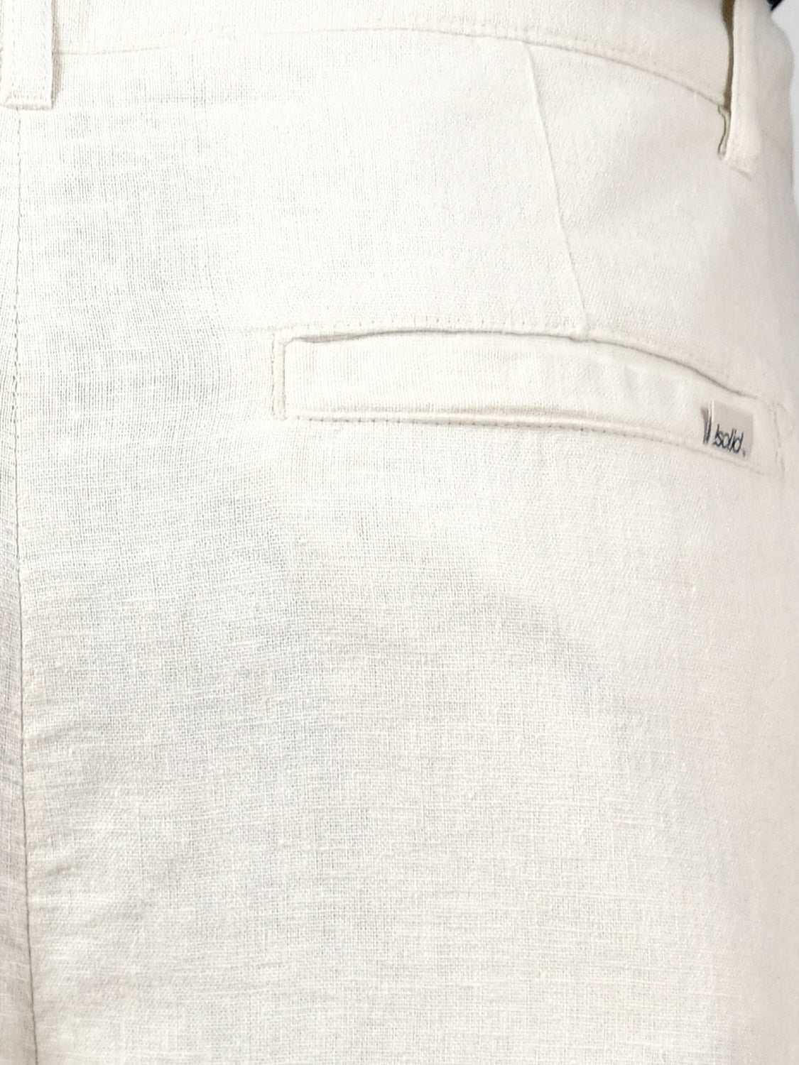 Close-up of the back pocket of the Ethan Tapered Pleat Pant by !Solid, featuring high-waisted, tapered pleated design in light beige and textured linen-viscose blend fabric with a small brand tag.