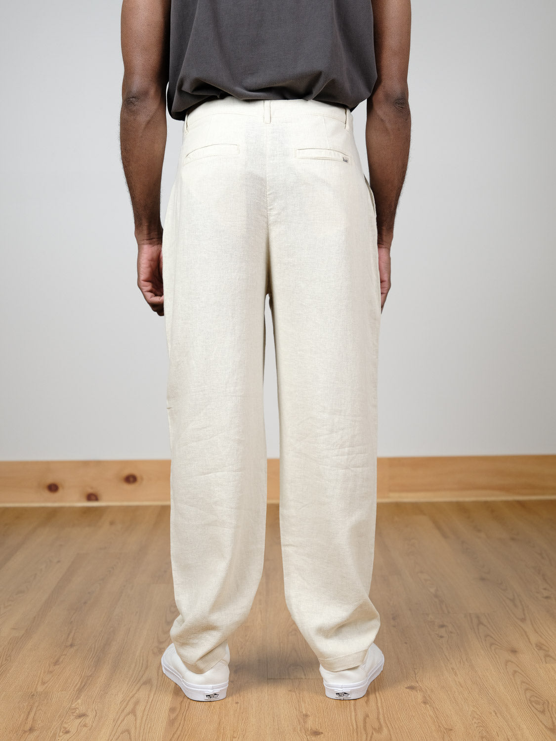 Shown from the waist down, a person stands indoors on a wooden floor wearing !Solid’s Ethan Tapered Pleat Pant in a cream linen-viscose blend, paired with a dark shirt and white sneakers against a plain light wall with wood baseboard.