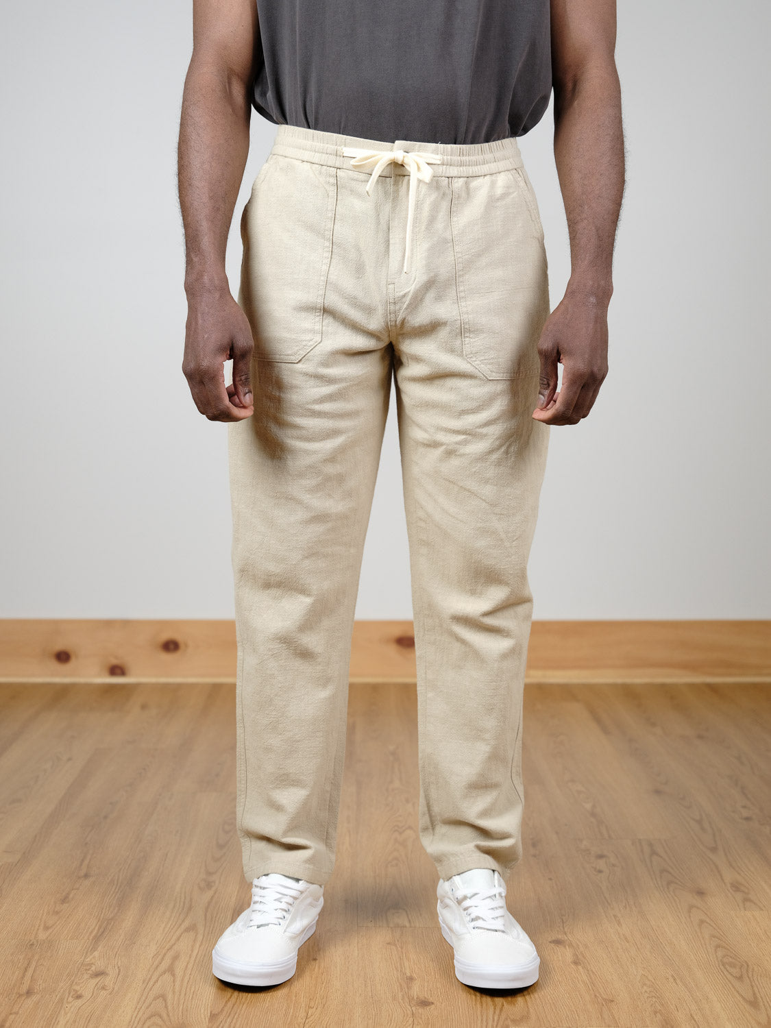 A person in ID Menswear's Sahara Linen Cotton Pant, a relaxed fit beige drawstring style, pairs them with a dark gray shirt and white sneakers while standing on a light wood floor against a plain light background.