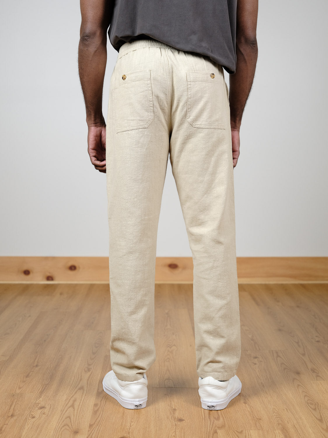 A person stands indoors on a wooden floor, facing away from the camera, wearing ID Menswear’s Sahara Linen Cotton Pant in beige, paired with white sneakers and a dark t-shirt.