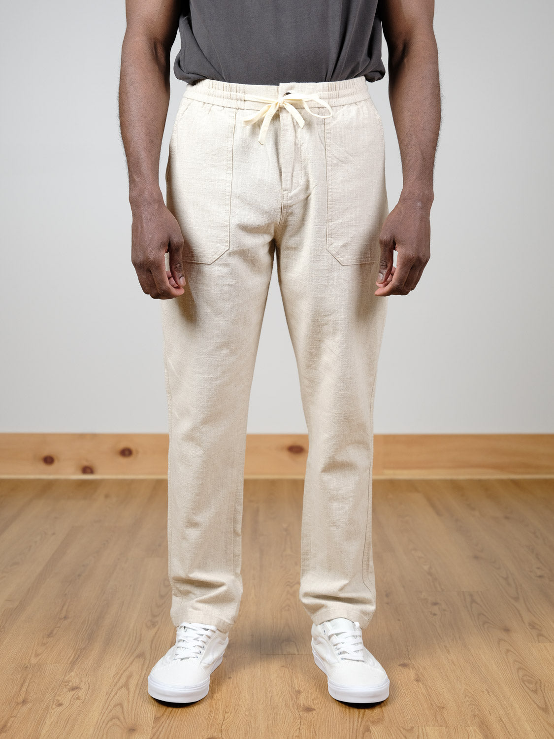 A person stands on a wooden floor wearing ID Menswear's Sahara Linen Cotton Pant in light beige, paired with a dark gray T-shirt and white sneakers; their head is not visible.