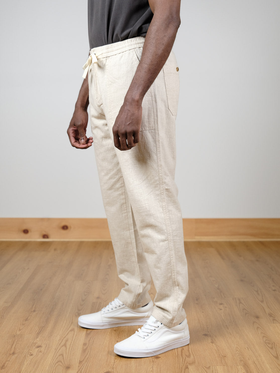Wearing the Sahara Linen Cotton Pant by ID Menswear in beige, paired with a dark tee and white sneakers, a person stands on a light wooden floor against a plain, light wall.