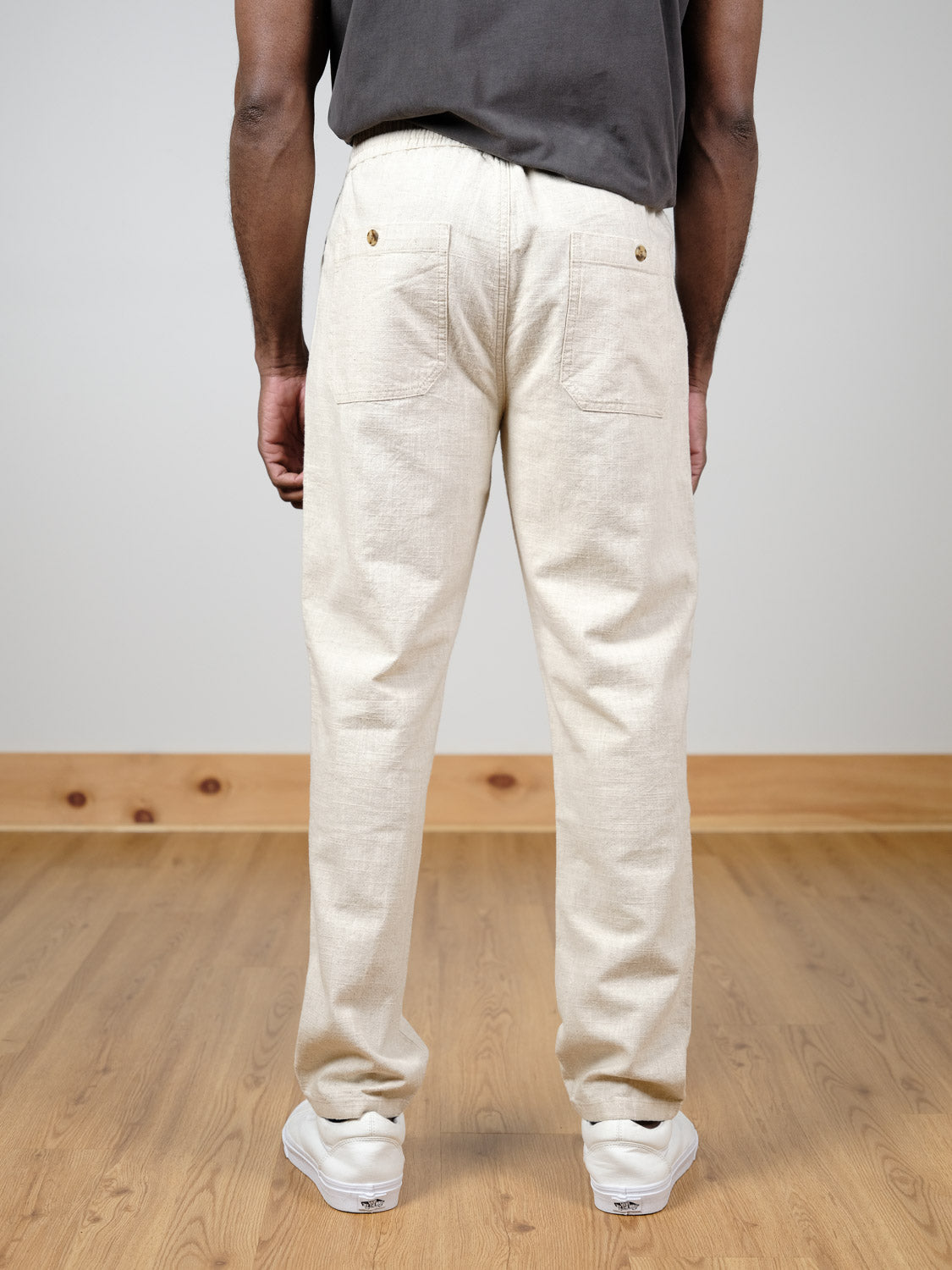 A person wears the ID Menswear Sahara Linen Cotton Pant in relaxed fit, light beige, with two buttoned back pockets and white sneakers, standing on a wooden floor facing away from the camera.