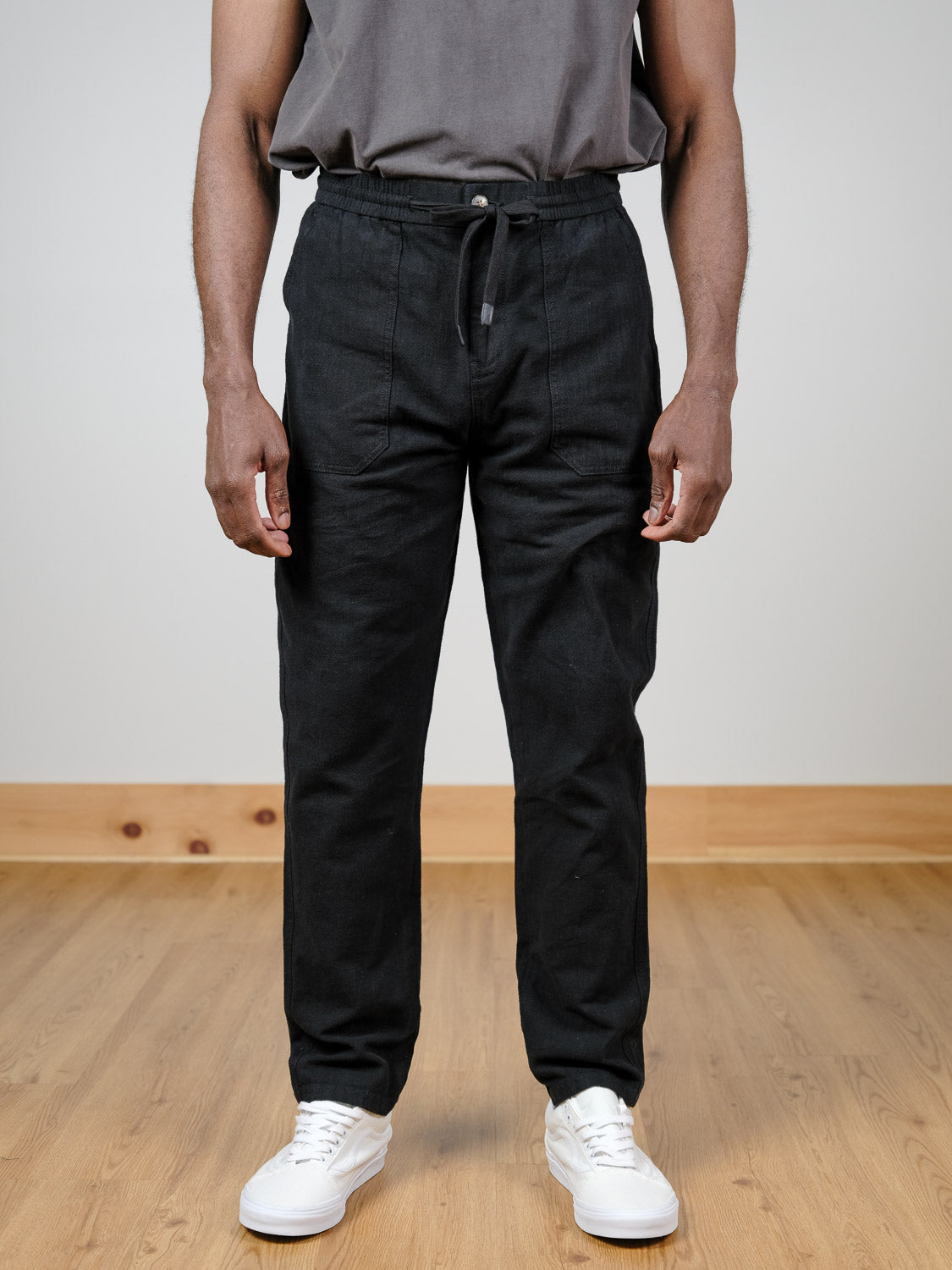 Indoors on a wooden floor, a person wears the Sahara Linen Cotton Pant by ID Menswear—black, relaxed fit, linen-cotton blend—with a gray t-shirt and white sneakers. Their face is not shown.