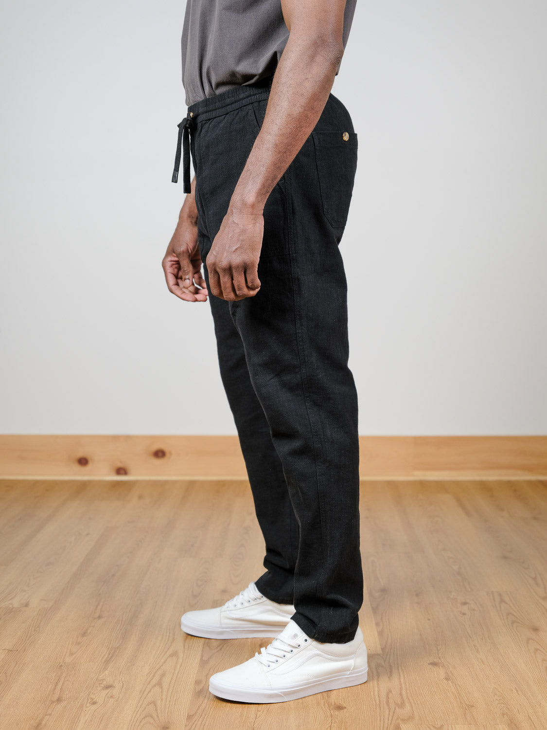 A person stands indoors on a wooden floor, wearing ID Menswear's Sahara Linen Cotton Pant in black, paired with a gray shirt and white sneakers. The side view shows the pants' relaxed fit from shoulder to feet, arms relaxed.