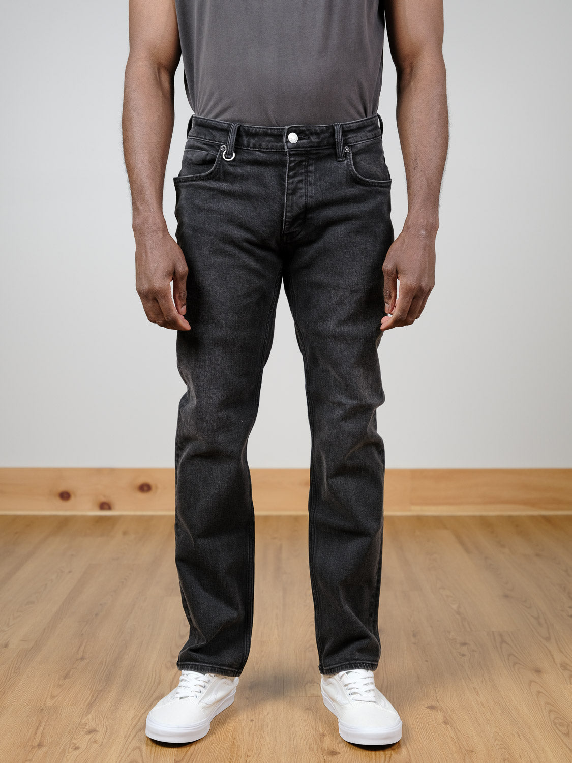 A person stands on a wooden floor against a plain light wall, wearing Neuw Lou Straight Cashbah jeans in faded black with a dark grey t-shirt and white sneakers. The photo is cropped from the shoulders down.