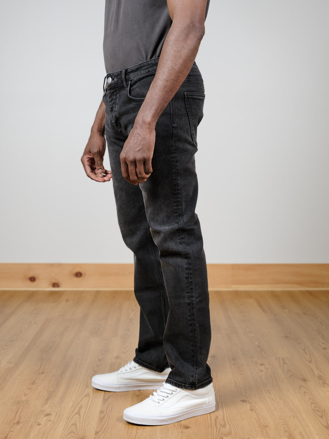 A person stands indoors on a wooden floor, wearing Neuw Lou Straight Cashbah jeans by Neuw in faded black denim, a dark gray t-shirt, and white sneakers. The photo is a side view from shoulders to feet against a plain light wall.