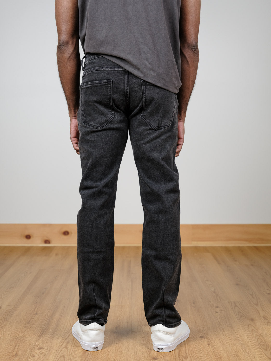 A person stands indoors on a wooden floor, wearing a dark t-shirt, white sneakers, and Neuw Lou Straight Cashbah jeans by Neuw in faded black denim. The setting features neutral-colored walls and a wooden baseboard.