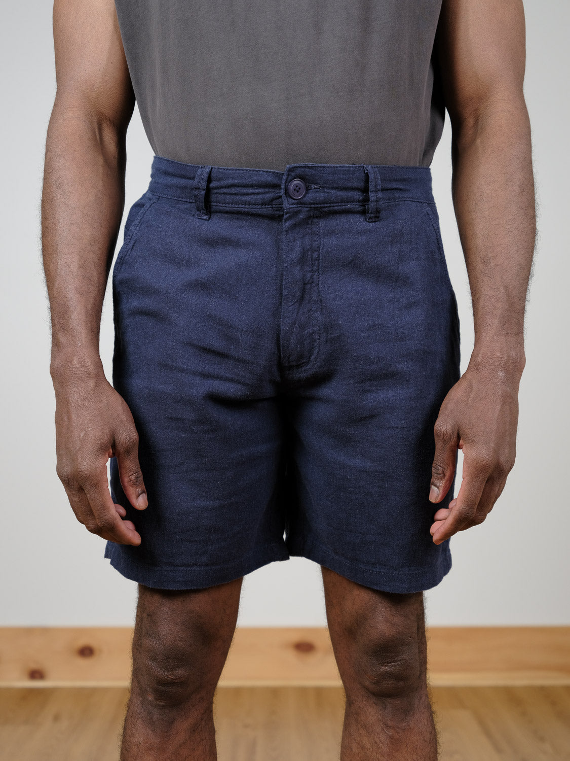 A person stands indoors wearing a grey shirt tucked into dark blue !Solid Aurelius Shorts. The image, cropped at the shoulders, shows their torso and legs against a plain light wall and wooden floor.