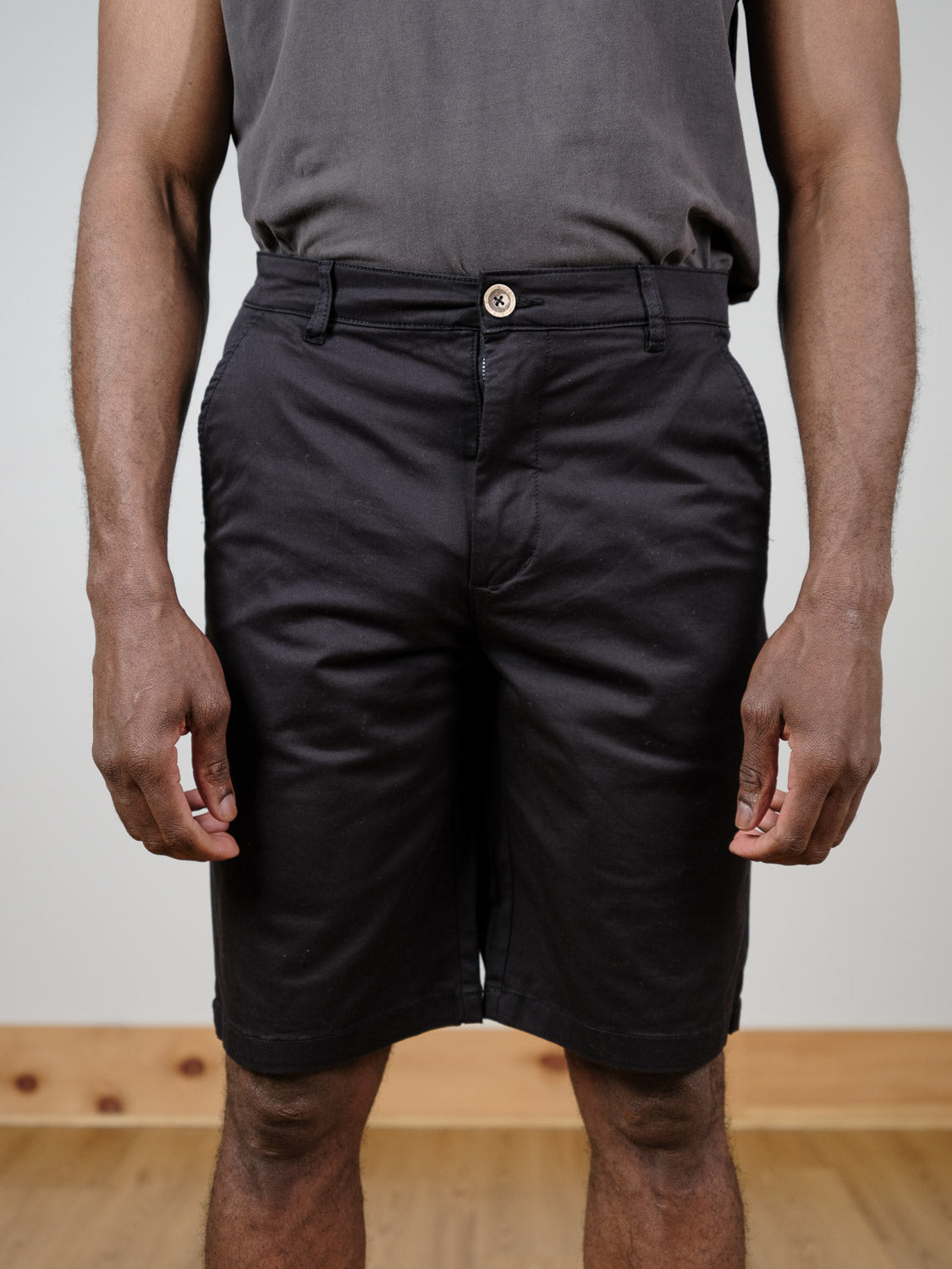 A person stands indoors wearing a dark gray t-shirt tucked into !Solid Bishop Shorts in black. Their hands rest relaxed at their sides; the wooden floor and baseboard are visible. The photo is cropped from shoulders to knees.