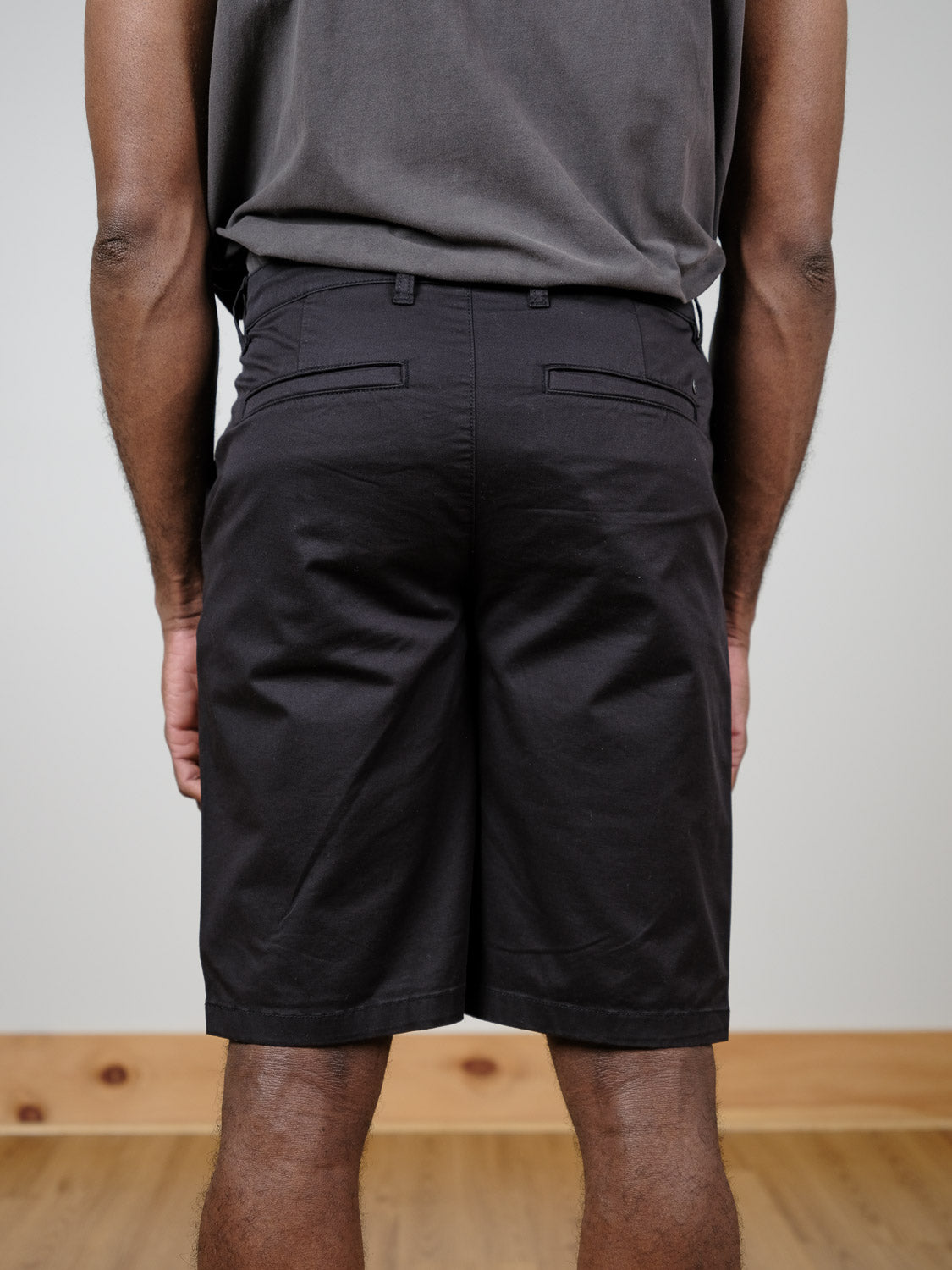 A person stands indoors facing away from the camera, wearing a dark sleeveless shirt and black !Solid Bishop Shorts; the background is a light-colored wall and wood floor.