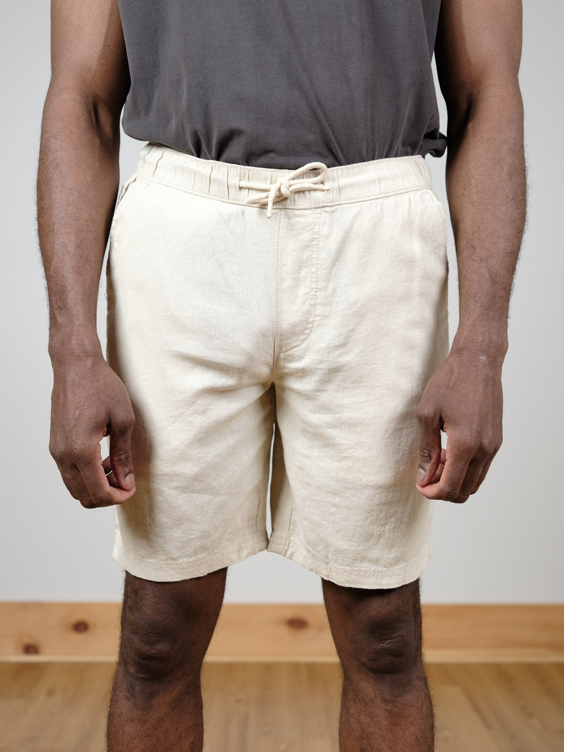 A person stands indoors on a wooden floor, arms relaxed by their sides, wearing light beige Aurelius Elas Shorts by !Solid and a dark gray t-shirt. The photo is cropped from shoulders to knees.