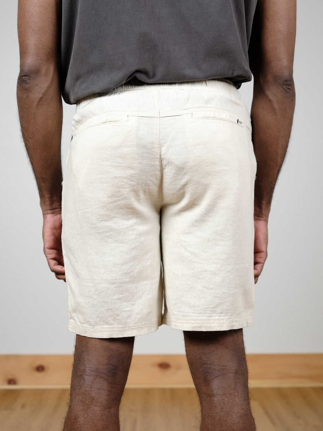 A person stands indoors with their back to the camera, wearing a dark shirt and light beige !Solid Aurelius Elas Shorts featuring a back pocket. Their arms are by their sides; the floor is wooden and the wall is plain.