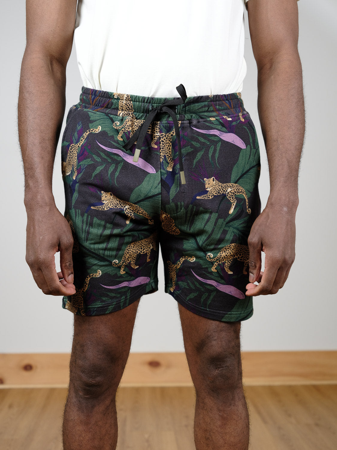 Someone stands indoors on a wooden floor wearing the Samui Leopard Print Short by ID Menswear, made from organic cotton with a drawstring and featuring a jungle leopard print and tropical leaf pattern.