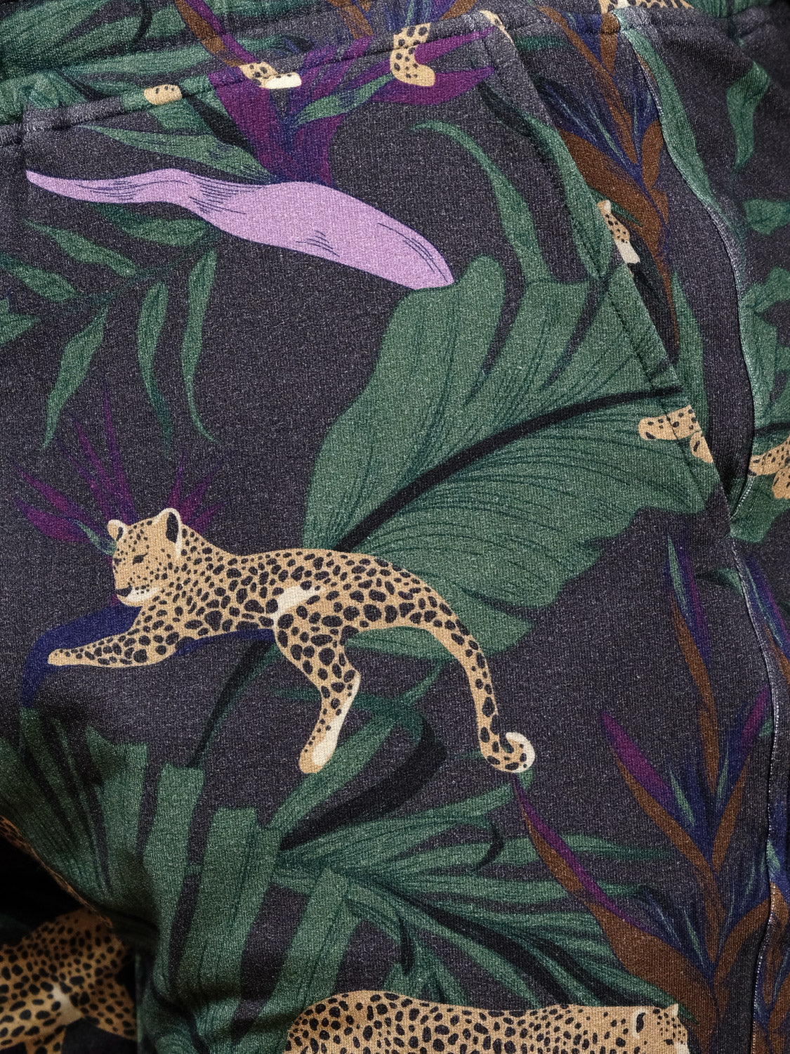 Close-up of the Samui Leopard Print Short by ID Menswear, featuring vibrant leopards, lush green leaves, and purple accents on a dark background—ideal for standout bamboo or organic cotton shorts.