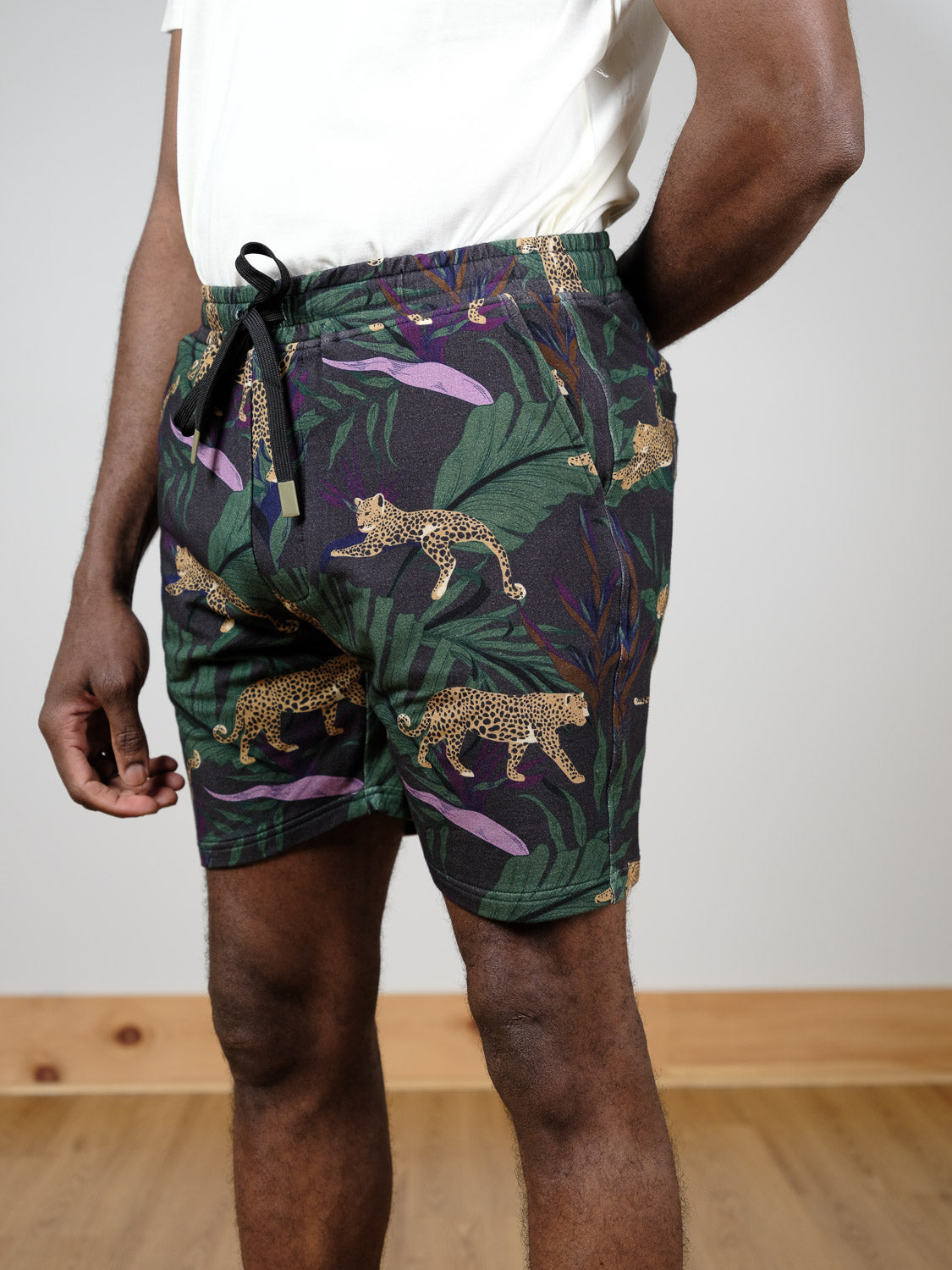 A person wears the Samui Leopard Print Short by ID Menswear, featuring a jungle leopard print with tropical leaf design, paired with a white shirt indoors on a wooden floor. Only the lower torso and upper legs are visible.