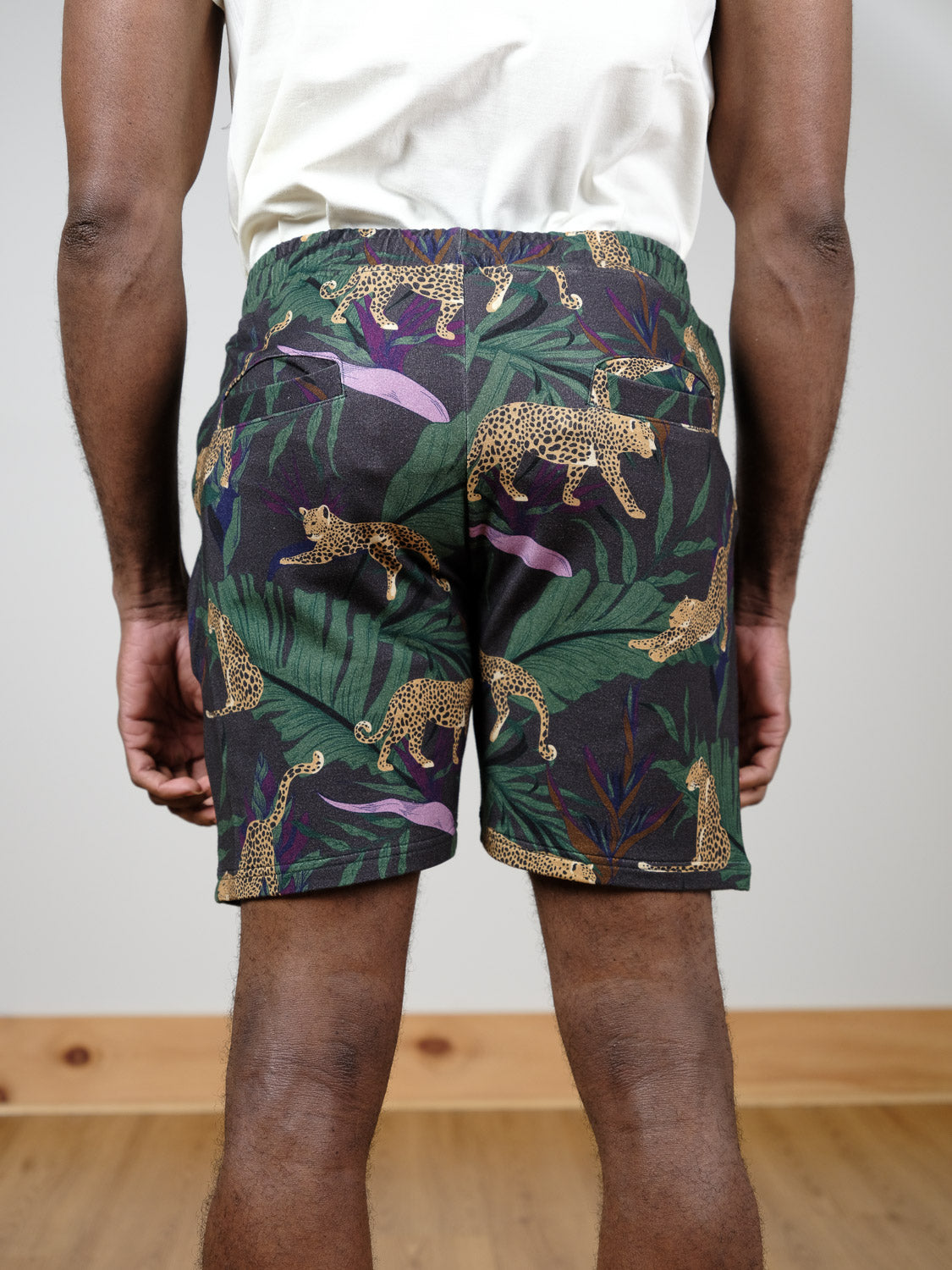 A person stands facing away, wearing the Samui Leopard Print Short by ID Menswear—organic cotton shorts with a jungle leopard pattern and green leaves. Their arms are relaxed at their sides on a wooden floor near a light-colored wall.