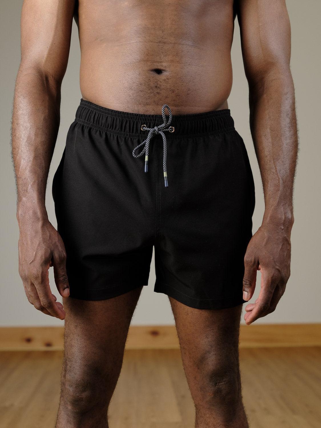 A man stands indoors on a wooden floor, wearing ID Menswear’s Oyogu Quick Dry 4 Way Stretch Swim Trunk in black, with his arms relaxed at his sides and only his torso and upper legs visible.
