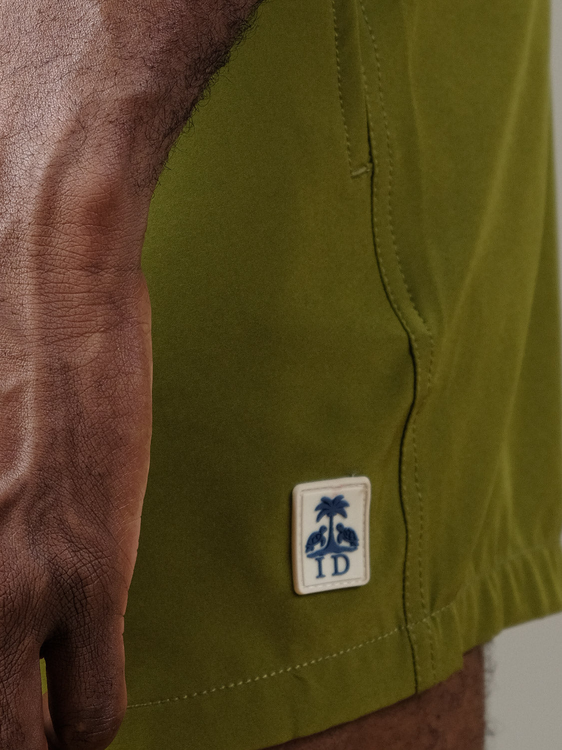 A close-up of a hand resting by the hip, wearing Oyogu Quick Dry 4 Way Stretch Swim Trunk by ID Menswear in olive green, featuring a stitched patch with two palm trees and the letters ID.