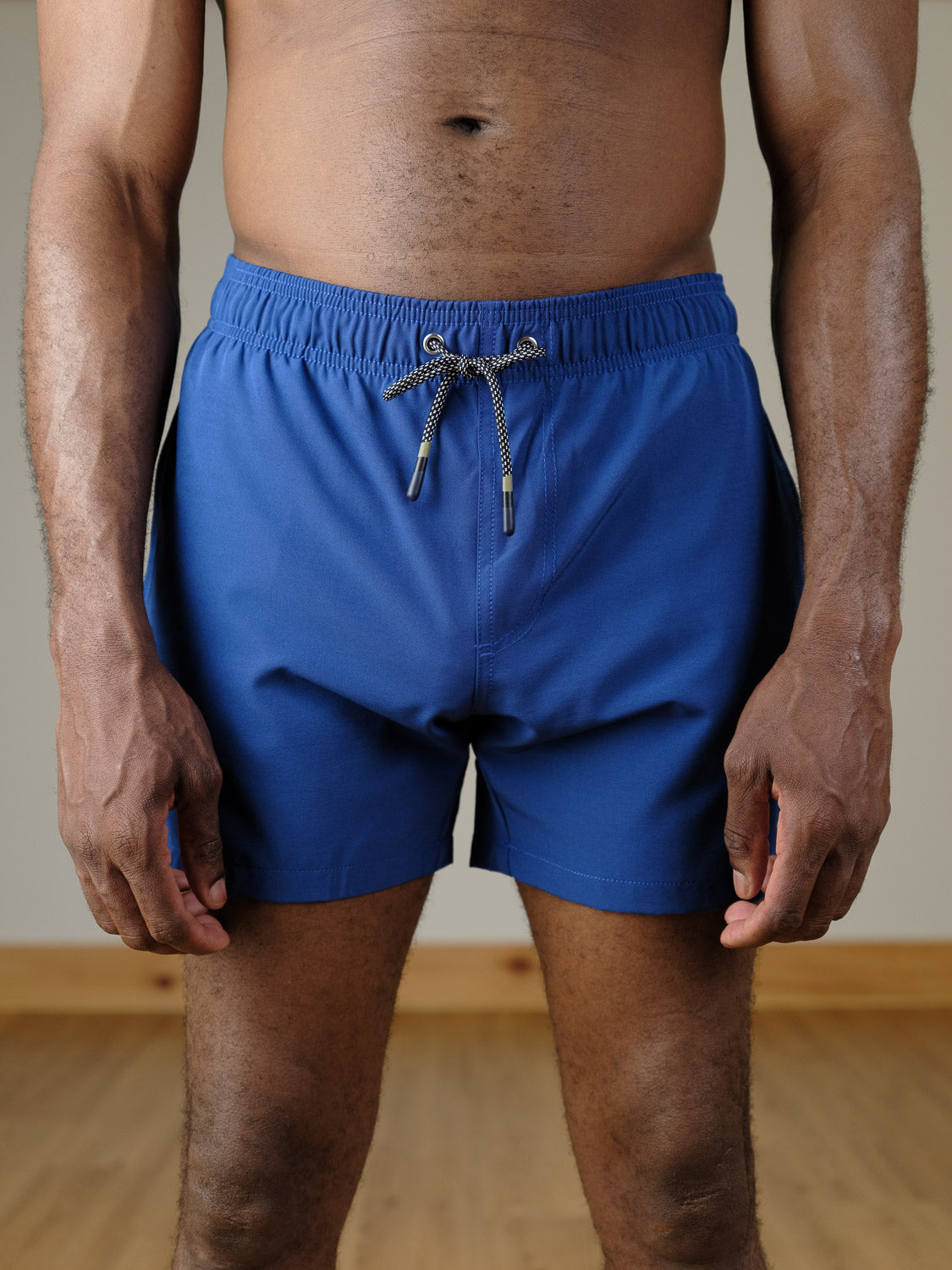 A person wearing Oyogu Quick Dry 4 Way Stretch Swim Trunk by ID Menswear stands indoors on a wooden floor, arms relaxed at their sides; the photo is cropped from shoulders to thighs.