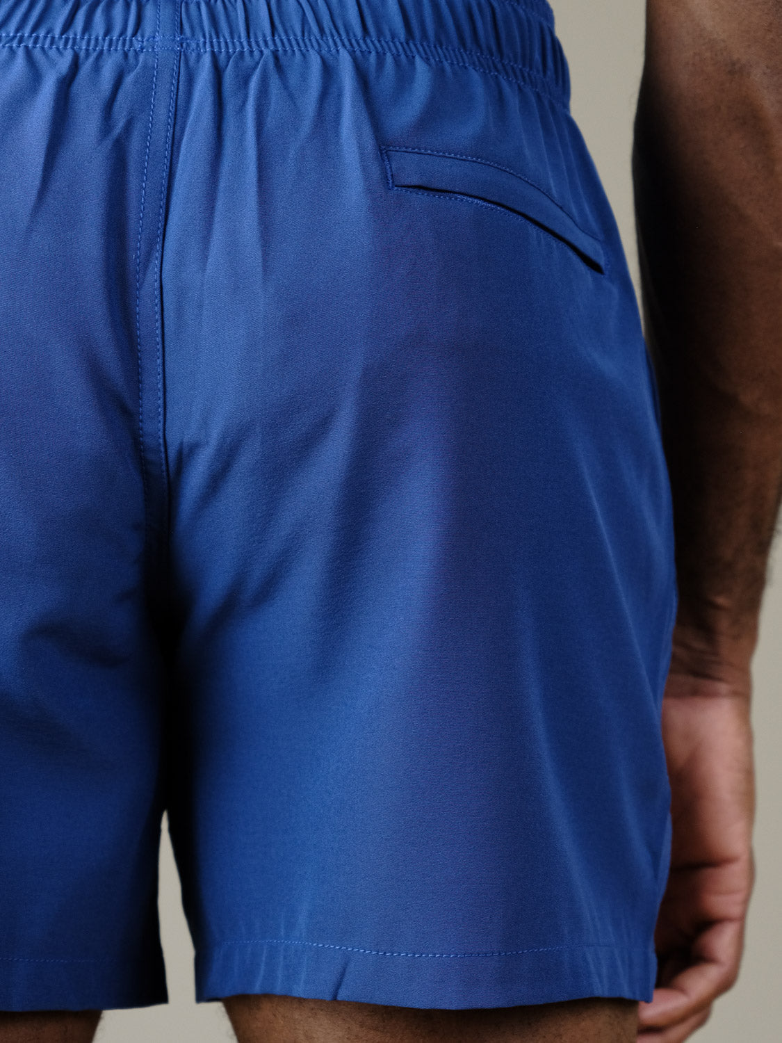 A close-up shows a person wearing ID Menswear’s Oyogu Quick Dry 4 Way Stretch Swim Trunk in blue, featuring an elastic waistband, a right-side back pocket, and part of their arm.