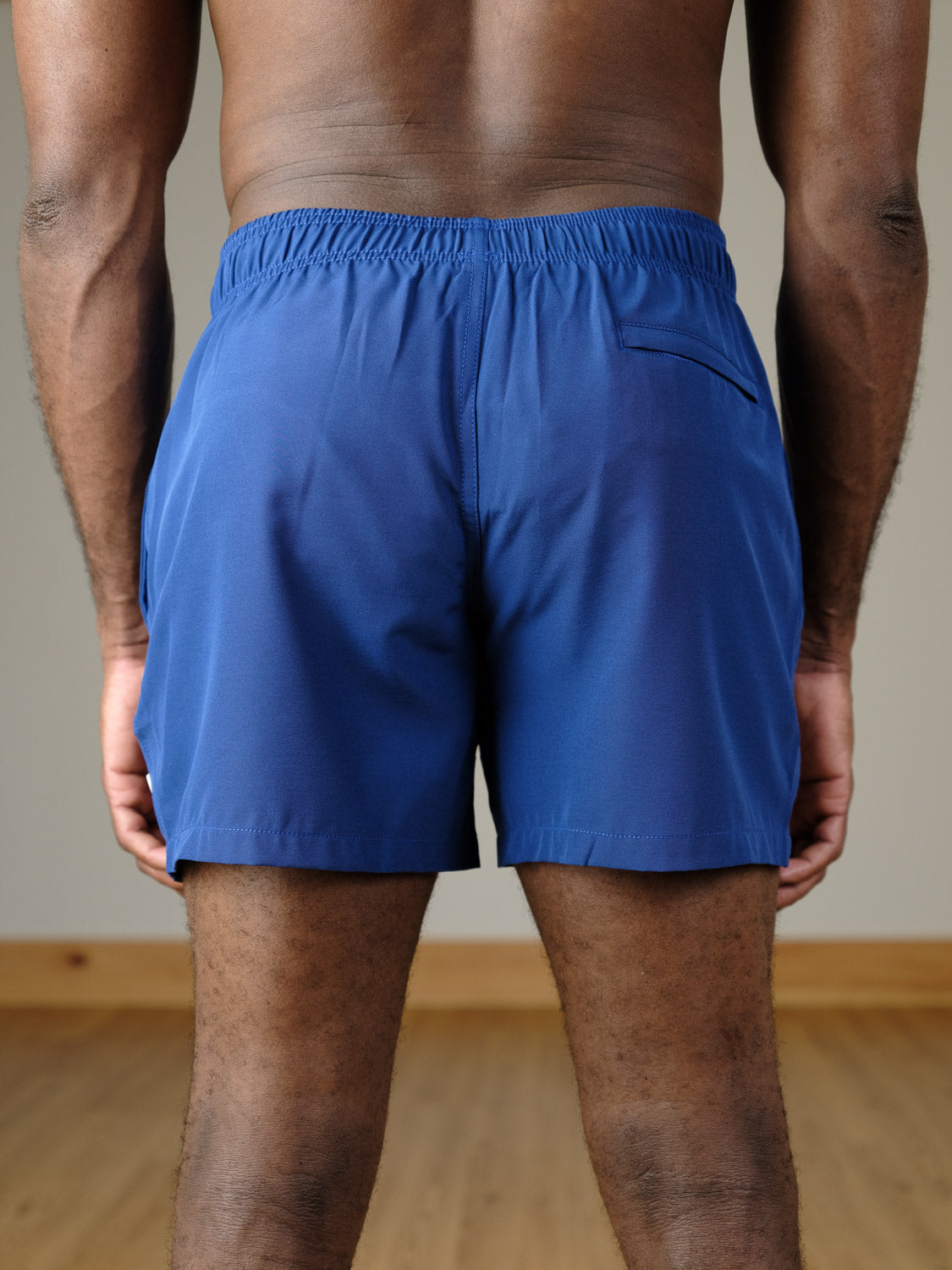 A person stands indoors on a wooden floor, facing away from the camera, wearing ID Menswear’s Oyogu Quick Dry 4 Way Stretch Swim Trunk in blue, featuring an elastic waistband and a small back pocket.