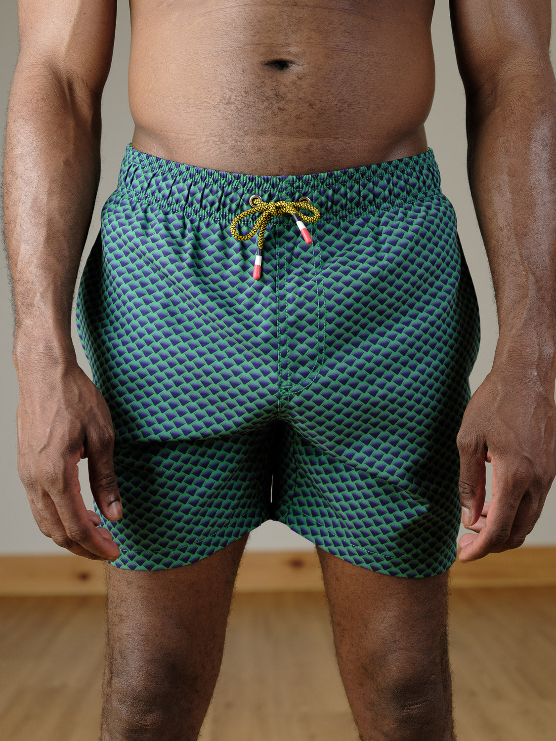 A person models the ID Menswear Uroko Fast Dry 4-Way Stretch Swim Trunk—patterned in green and blue—indoors on a wooden floor, shown from shoulders down with arms relaxed at their sides.