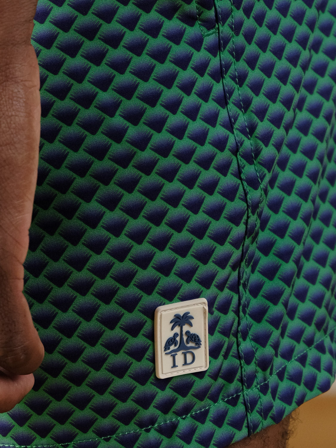 Close-up of blue and green textured fabric from the Uroko Fast Dry 4-Way Stretch Swim Trunk by ID Menswear, featuring a small patch with palm trees, turtles, and “ID.” Part of a hand and leg appear at the image’s edge.