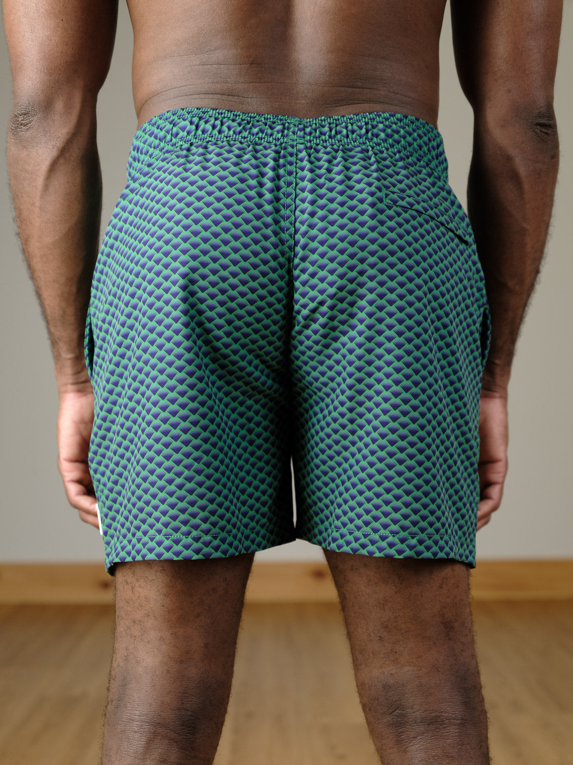 A person stands indoors facing away, wearing ID Menswear's Uroko Fast Dry 4-Way Stretch Swim Trunk in a green and blue pattern with an elastic waistband. The background has a plain wall and wooden floor.