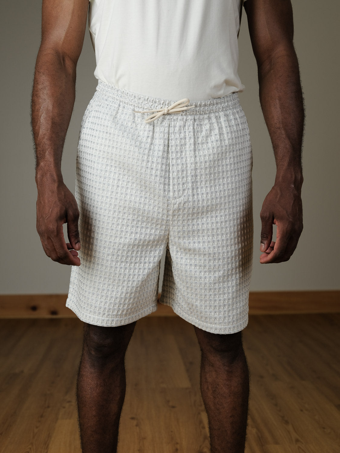 A person wearing Les Deux's Check Seersucker Shorts and a white textured sleeveless shirt stands on a wooden floor with their arms relaxed at their sides.