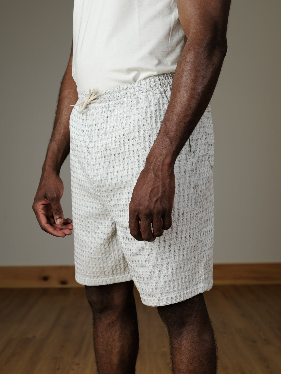A person wearing Les Deux Check Seersucker Shorts stands indoors on a wooden floor, with only their torso and legs visible.