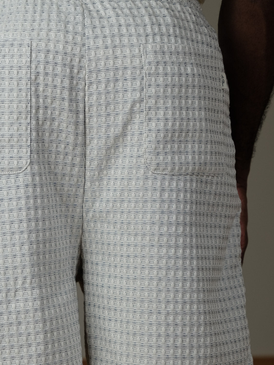 Close-up of the back of a person wearing Les Deux Check Seersucker Shorts, featuring an elastic waistband, two rear pockets, and a light-colored square grid pattern. The person's arm is partially visible on the right.