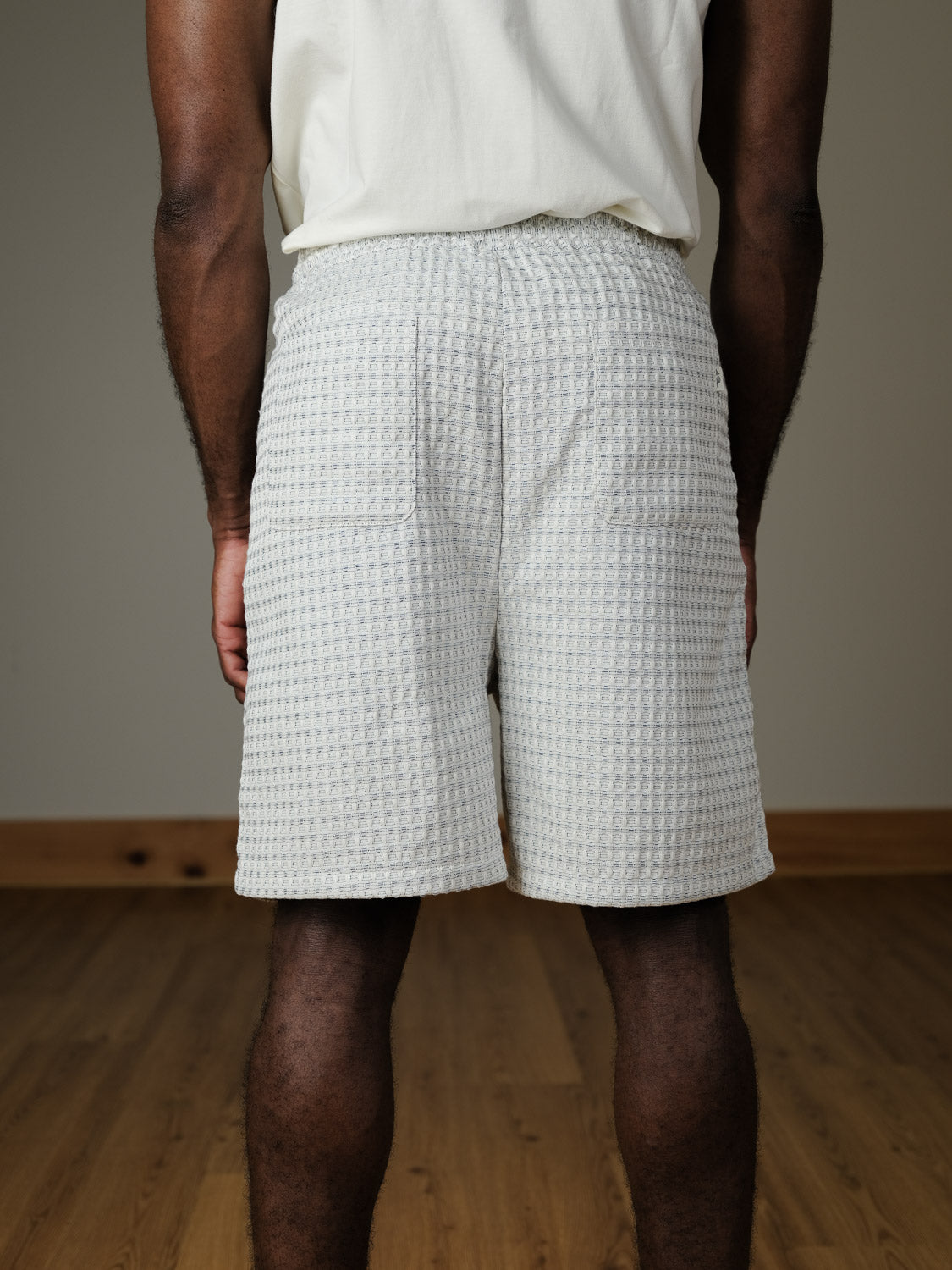 A person stands indoors on a wooden floor, shown from the back, wearing a white shirt and Les Deux Check Seersucker Shorts—textured white shorts featuring two back pockets.